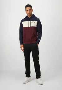 Maroon and navy color-blocked hoodie with white chest section, featuring a front pocket and drawstrings. Paired with black pants and white sneakers.