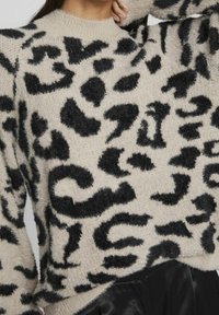 Beiger Pullover mit schwarzen, flauschigen Leopardenmustern. Rundhalsausschnitt, lange Ärmel, weicher Stoff und eine leicht verkürzte Passform.