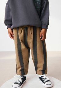 Bambino che indossa pantaloni marroni oversized con strisce verticali nere, una felpa grigia scura e sneakers nere con lacci bianchi.