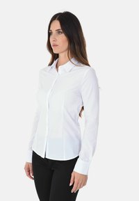 Camicetta bianca a maniche lunghe con colletto a punta, vestibilità strutturata, texture liscia e orlo curvo. Indossata con pantaloni neri.
