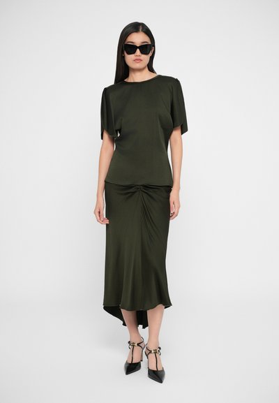 Abito in satin verde oliva con dettaglio drappeggiato sul davanti, maniche corte e orlo asimmetrico. Abbinate a tacchi neri a punta e occhiali da sole.