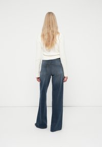 Žena s dlhými blond vlasmi, ktorá má na sebe biely pletený sveter a modré nohavice širokého strihu, stojí čelom k bielej stene.