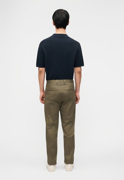 Άνδρας που στέκεται με την πλάτη του, φοράει μπλε ναυτικό υφασμάτινο κοντομάνικο πουκάμισο, ελιάς πράσινο παντελόνι και λευκά αθλητικά παπούτσια μπροστά σε απλό φόντο.