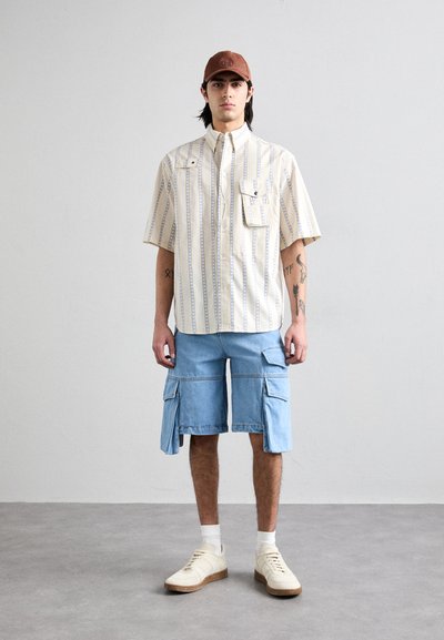 Camicia a maniche corte a righe in crema e blu con taschino, abbinata a pantaloni cargo azzurri chiari con tasche laterali e sneakers beige.
