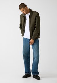 Olivgrüne Bomberjacke mit Kragen, kombiniert mit weißem T-Shirt und hellblauen Jeans, ergänzt durch schwarze Schuhe.