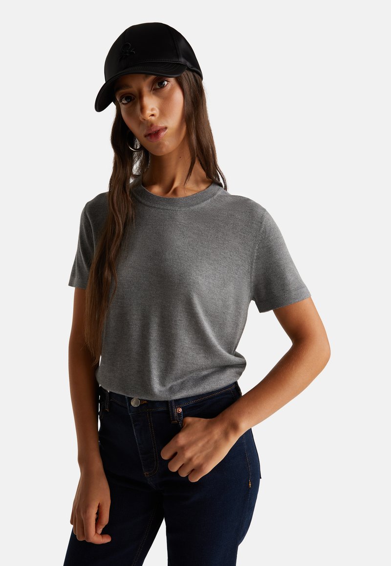 Šedé tričko s krátkým rukávem a kulatým výstřihem, spojené s tmavě modrými džínami. Model nosí černou čepici s jemným logem.