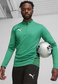 Groene, lange mouw sporttop met een halve ritssluiting en Puma-logo, gecombineerd met zwarte shorts met een logo. Persoon houdt een voetbal vast.