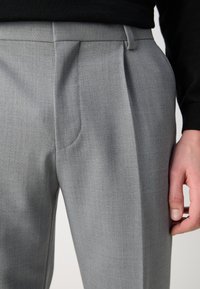 Lysegrå skreddersydde bukser med en teksturert stoff, med legg ved livet og en glatt finish, vist med en svart topp som delvis er synlig.