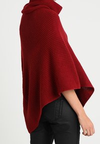Personne portant un poncho en tricot rouge à col haut sur un pantalon noir, vue de profil sur un fond uni.