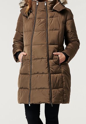 Femme portant un manteau d'hiver matelassé marron longueur genou avec une capuche bordée de fourrure et deux poches avant zippées, mains dans les poches.