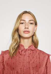 Blusa rosa con lunares texturizados, hombros con volantes y un lazo en el cuello. Tejido ligero y transparente con rayas verticales y un suave brillo.
