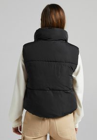 Black puffer vest with high collar, quilted design, and a smooth texture, worn over a light-colored long-sleeve shirt and beige pants.
