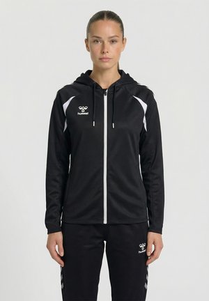 Femme debout face à l'avant portant une veste à capuche noire zippée et un pantalon assorti avec des logos et bandes blanches Hummel.