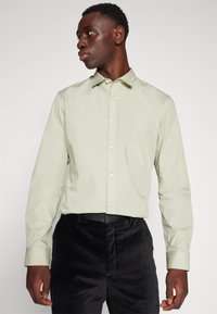 Chemise à manches longues vert clair avec bouton devant, col soigné, texture subtile, associée à un pantalon noir en tissu doux.