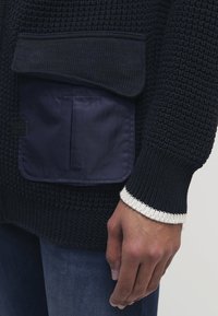 Närbild av en hand bredvid en mörk stickad tröja med en stor ficka i marinblått tyg och detalj på ärmarna över blå jeans.