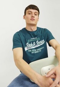 Tyrkisk bomulds T-shirt med hvid grafisk tekst, rund halsudskæring og korte ærmer, båret med blå denimjeans og en lysefarvet taske.