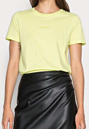 Frau trägt ein hellgelbes, kurzärmeliges T-Shirt, das in einen hoch taillierten schwarzen Lederrock mit seitlichem Drapée-Detail gesteckt ist.