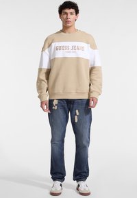 Beżowa i biała bluza z logo "GUESS JEANS", w połączeniu z niebieskimi przetartymi dżinsami i białymi trampkami. Miękki materiał bawełniany, luźny krój.