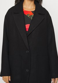 Manteau en mélange de laine noir avec un col noté, deux poches avant et une fermeture à deux boutons. Le tissu a une texture lisse.