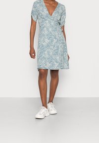 Blå omslagklänning med vitt blommönster, korta ärmar, v-ringning och knuten midja, i kombination med vita sneakers mot en neutral bakgrund.