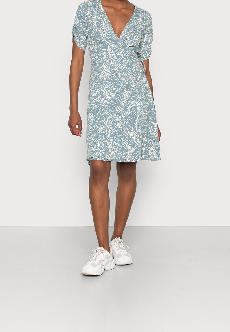 Blå omslagklänning med vitt blommönster, korta ärmar, v-ringning och knuten midja, i kombination med vita sneakers mot en neutral bakgrund.