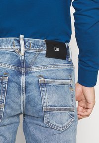 Riflové džíny v světle modré barvě s černou štítkem LTB na pásku a kontrastním žlutým šitím. Zadní pohled ukazuje dvě zadní kapsy.