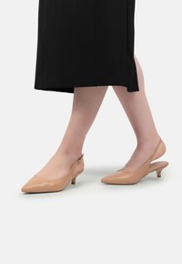 Sandalete bej cu toc slingback, vârf ascuțit, toc mic tip bobină și finisaj lucios din patent. Designul include o decupare mare în partea din spate.