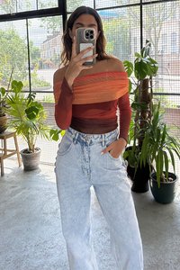 Off-shoulder fargeblokkert topp i nyanser av rød og oransje, kombinert med høyhåndt høyvasket jeans. Innendørs med grøntområder.