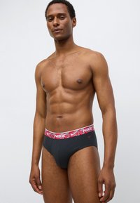 Černé slipy Nike s červenobílým vzorovaným pasem s logem Nike. Vyrobené z hladké látky, přiléhavý střih, bez výrazných ozdob.