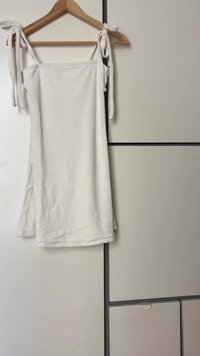 Vestido blanco sin mangas con tirantes de lazo, confeccionado en una tela suave, con un corte holgado y colgado de una percha de madera sobre un fondo claro.