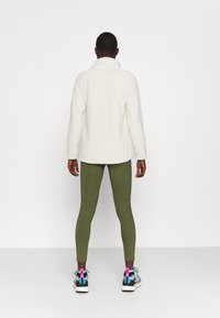 Weißer Fleece-Pullover mit hohem Kragen, kombiniert mit olivgrünen Leggings und bunten, gemusterten Sneakers mit einer dicken Sohle.