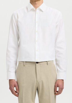 Homme portant une chemise blanche unie à manches longues rentrée dans un pantalon beige, sur un fond clair.