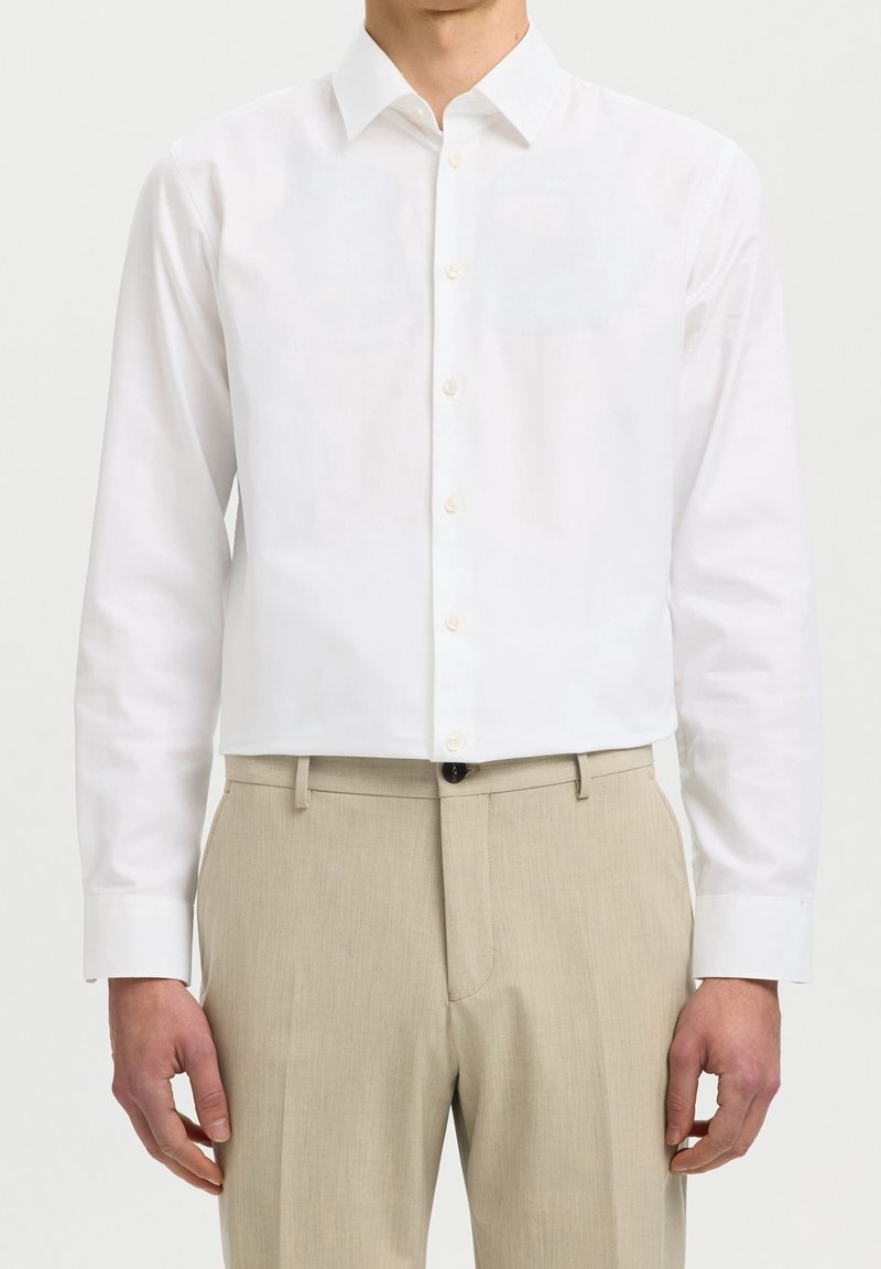 Homme portant une chemise blanche unie à manches longues rentrée dans un pantalon beige, sur un fond clair.