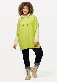 Pullover oversize verde lime con cappuccio, abbinato a una maglia a maniche lunghe a righe e jeans in denim scuro, con stivali beige con lacci.
