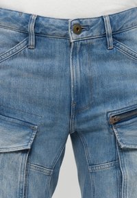 Blue denim cargo pants with zipper pocket on right thigh and button closure at waist, showing detailed stitching and fabric texture.