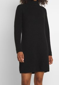 Robe à col roulé en tricot noir avec des manches longues, longueur mi-cuisse et texture côtelée. Présente des fentes sur les côtés pour faciliter les mouvements.