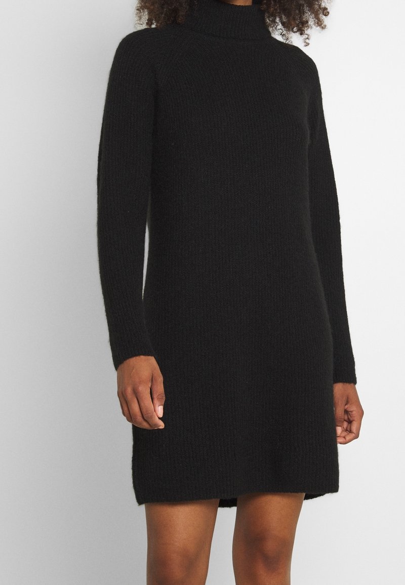 Robe à col roulé en tricot noir avec des manches longues, longueur mi-cuisse et texture côtelée. Présente des fentes sur les côtés pour faciliter les mouvements.