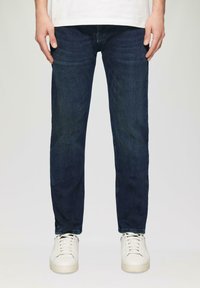 Jean en denim bleu foncé avec une coupe droite, présentant un délavage subtil et des marques de frottement, associés à des baskets blanches.