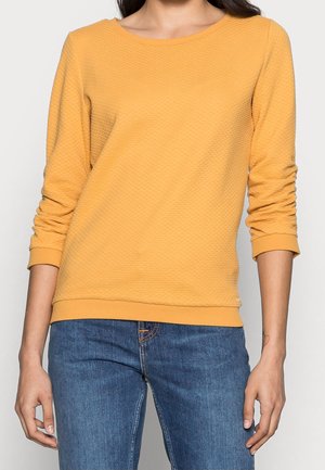 Persoon draagt een mosterdgele longsleeve met tekstuur en geplooide mouwen, en een blauwe spijkerbroek, staat met ontspannen armen.