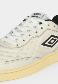 Λευκό και μπεζ αθλητικό παπούτσι Umbro με μαύρο λογότυπο στο πλάι και τη γλώσσα, λευκά κορδόνια και μπεζ σόλα.