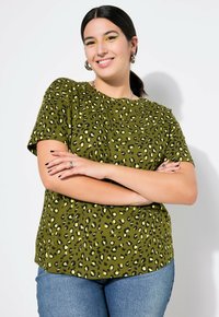 Olivgrünes Shirt mit einem schwarz-gelben Leopardenmuster, kurzen Ärmeln, Rundhalsausschnitt und lockerer Passform. Wird mit blauen Jeans getragen.