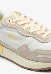 Sneaker met een beige suede bovenkant, witte textile accenten, een crèmekleurige rubberen zool, gele veters en een gele krokodillen logo detail.