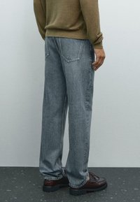 Persona che indossa jeans azzurri a gamba dritta, scarpe di pelle marrone e un maglione verde oliva, in piedi su un pavimento scuro con macchie.