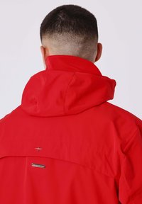 Man met kort donker haar, in een helderrode jas met een opstaande kraag, gezien van achteren tegen een eenvoudige lichte achtergrond.