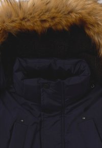 Marineblauwe pufferjas met een grote capuchon van nepbont. Voorzien van een hoge kraag, drukknoopsluiting en een zachte, pluizige binnenvoering.