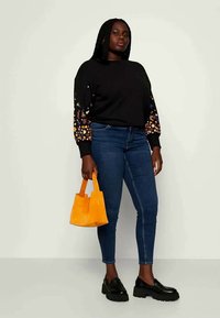 Felpa nera con maniche ricamate a fiori, jeans skinny blu, scarpe con tacco nere e una piccola borsa arancione.