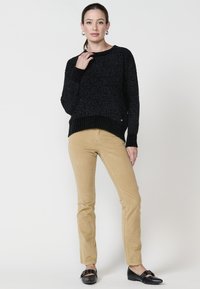 Črna teksturirana pletena jopica z rebrastim robom, kombinirana z bež kordnimi hlačami. Manekenka nosi črne čevlje brez vezalk in pozira z eno roko na ovratniku.