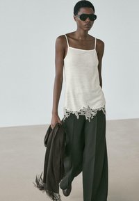 Frau mit schwarzen Sonnenbrillen, ärmellosem weißen Oberteil mit Spitzenabschluss, weiten schwarzen Hosen, hält einen dunkel gefransten Schal und geht drinnen.