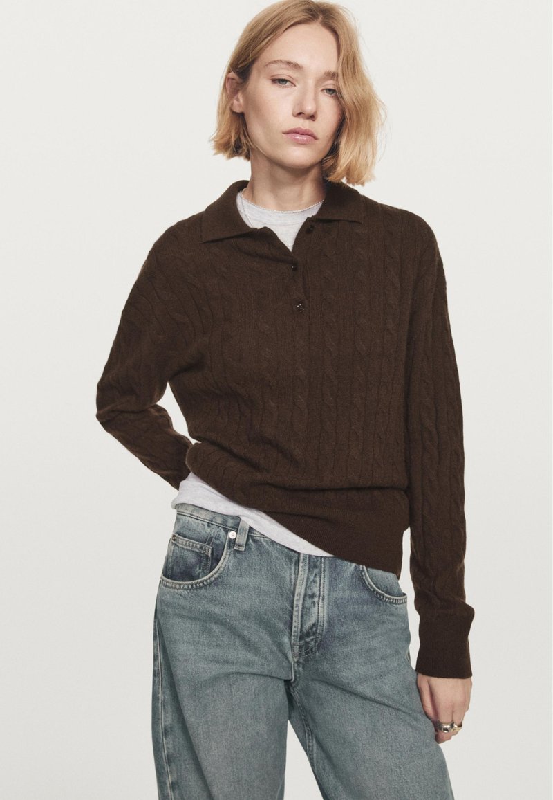 Giovane con capelli biondi corti che indossa un maglione con colletto a trecce marrone scuro sopra una camicia chiara e jeans blu larghi, su uno sfondo bianco.