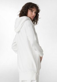 Sudadera blanca con capucha y mangas largas, que presenta un bolsillo tipo canguro, puños de canalé y una textura suave. Vista trasera con un corte relajado.